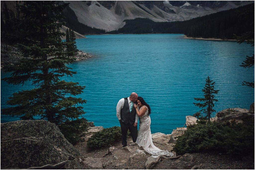 Bride and groom intimately embracing on some rocks overlooking a perfectly aquamarine Moraine Lake in Banff National Park