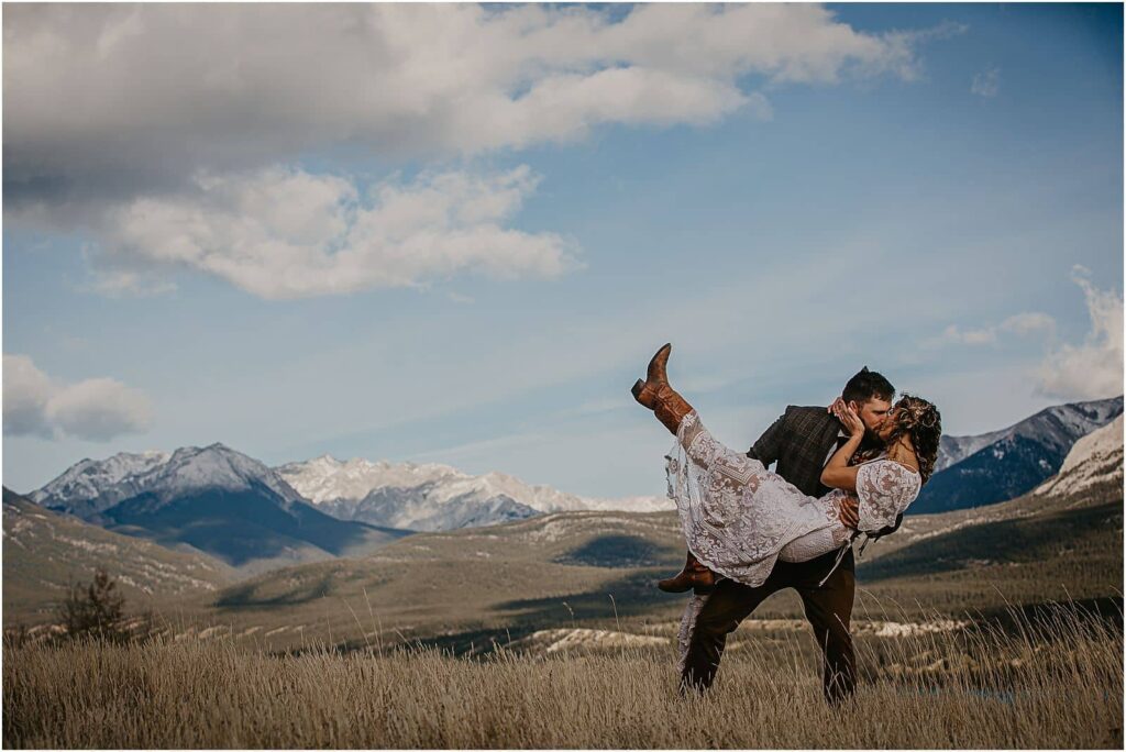 A groom holding his bride in a bridal lift against a mountain backdrop while dipping her and they are kissing