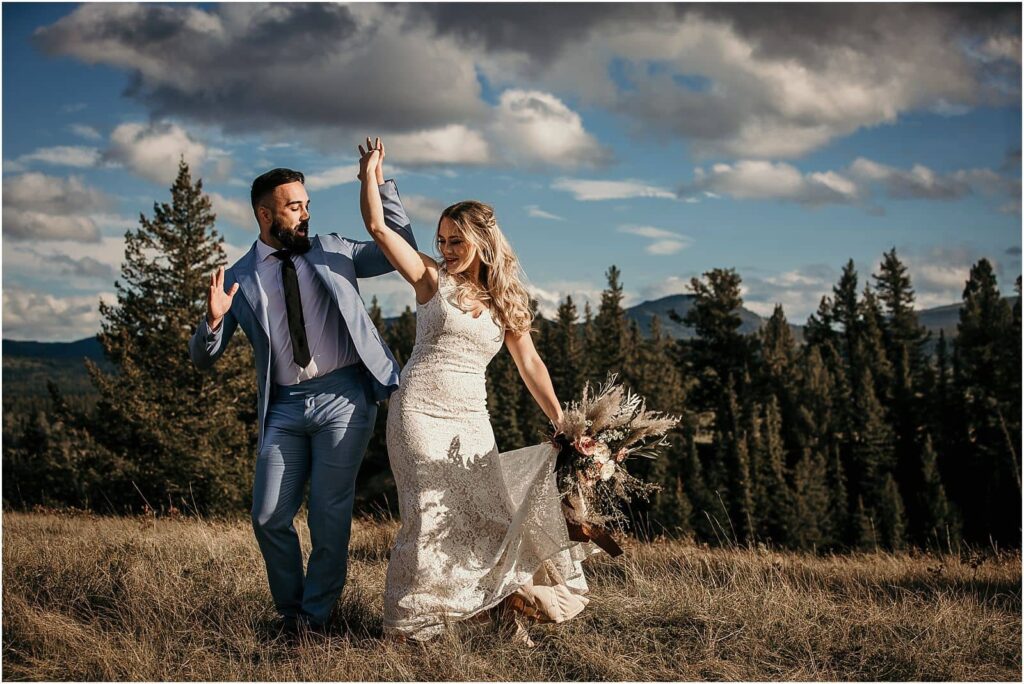 A bride and groom hip-bumping and smiling with their arms raised in a hand-hold above their heads with mountains and trees in the background