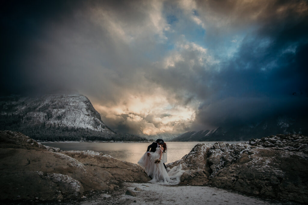 Couple eloping at Lake Minnewanka during a winter sunrise with snow-covered mountains and soft morning light.