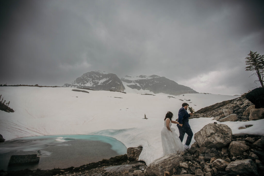 Couple atop a mountain via helicopter in Revelstoke beside a turquoise glacier pool during an adventure elopement.