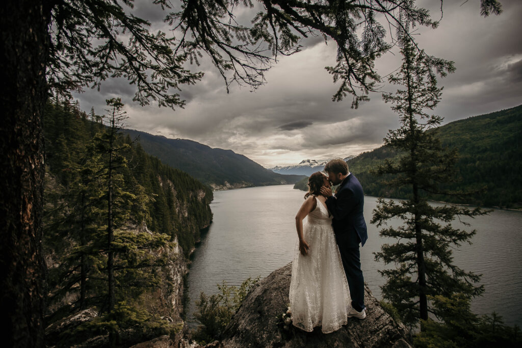 Couple eloping on a cliff in Revelstoke overlooking a deep blue lake and surrounding mountains.